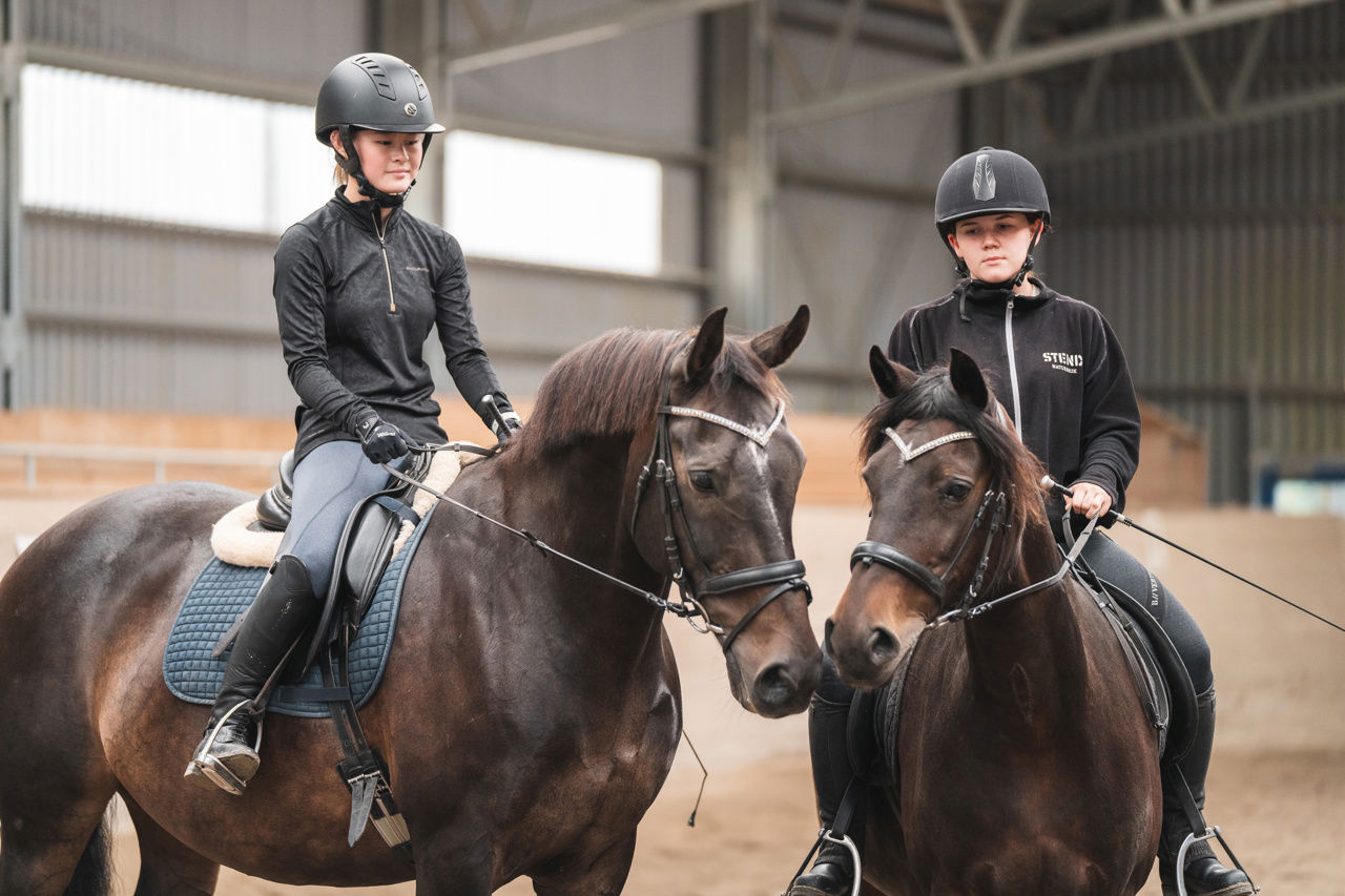 Bildet viser to jenter som sitter på hver sin brun hest i ridehallen