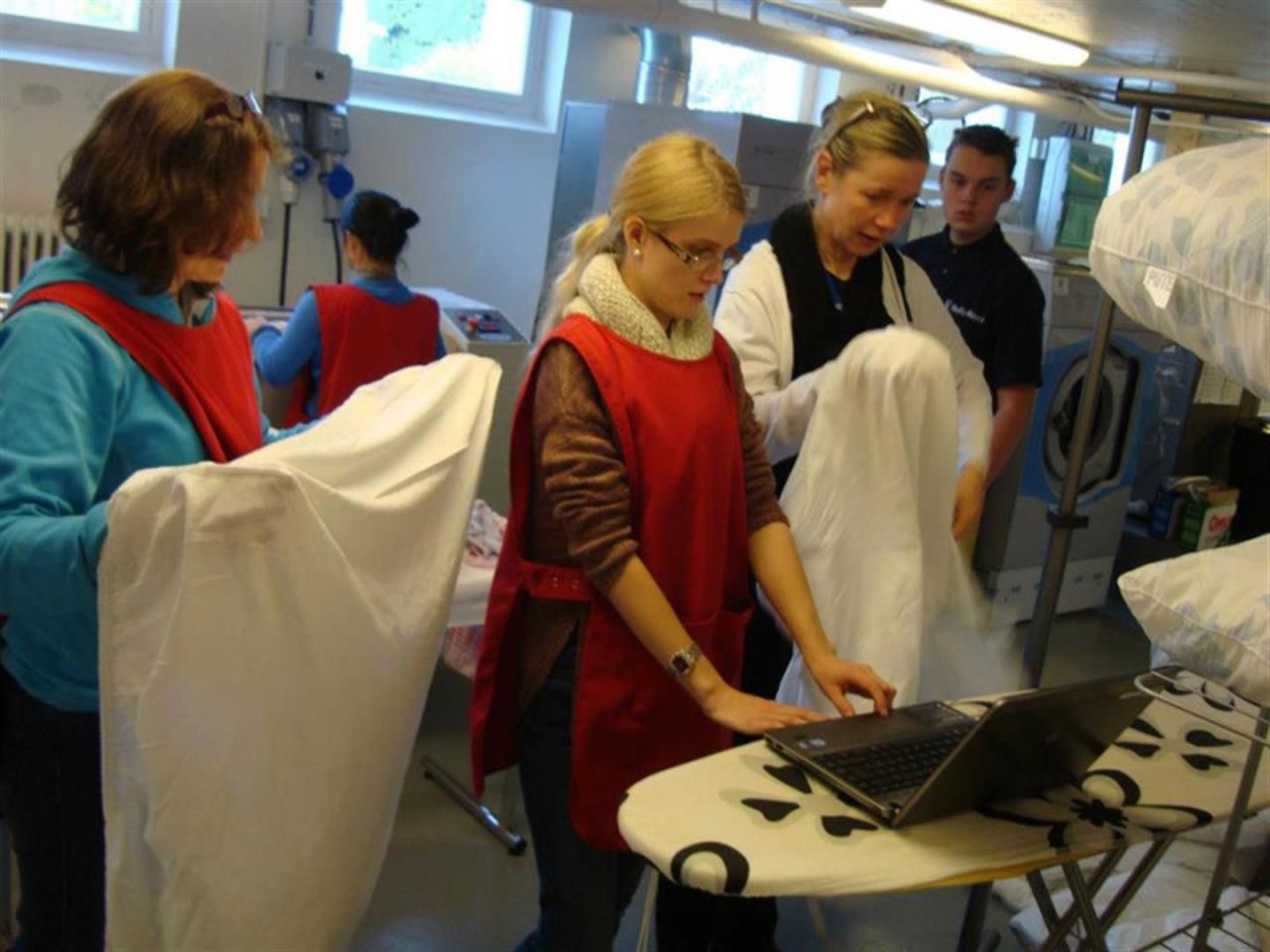 Bildet viser elever og lærere som bretter stretcs-laken på vaskeri og ser på en PC